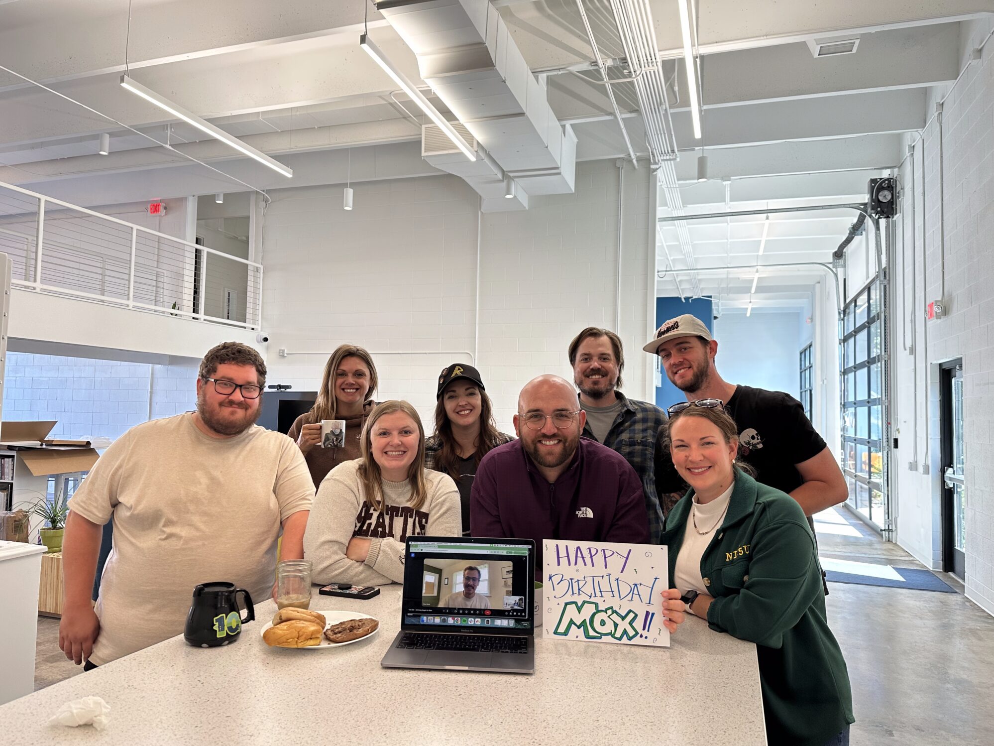 A group of eight people gather around a kitchen island for a photo. There is also someone joining virtually from a laptop. There are bagels on a plate on the island, along with a small sign that reads “Happy Birthday Max!”