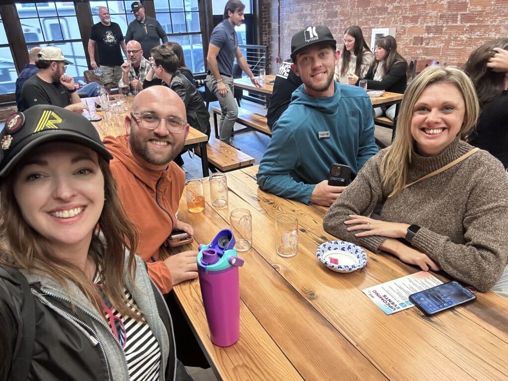 Four people sit at a long wooden table in a busy restaurant or brewery, smiling at the camera. The woman on the left wears a black hat with pins and a striped shirt, next to a man in glasses and an orange hoodie. Across from them are a man in a teal sweatshirt and black cap holding a phone, and a woman in a brown sweater with a smartwatch. Empty pint glasses, a colorful water bottle, and a phone rest on the table. In the background, other groups of people are gathered around tables against brick walls and large windows.