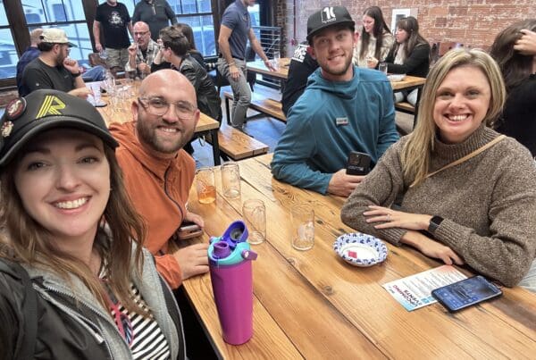 Four people sit at a long wooden table in a busy restaurant or brewery, smiling at the camera. The woman on the left wears a black hat with pins and a striped shirt, next to a man in glasses and an orange hoodie. Across from them are a man in a teal sweatshirt and black cap holding a phone, and a woman in a brown sweater with a smartwatch. Empty pint glasses, a colorful water bottle, and a phone rest on the table. In the background, other groups of people are gathered around tables against brick walls and large windows.