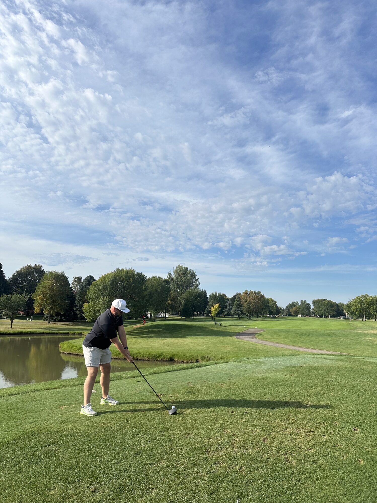 A man wearing a white hat, black polo, and khaki shorts holds a gold club. He is preparing to hit the golf ball. The grass is green and the sky is bright blue with few clouds.