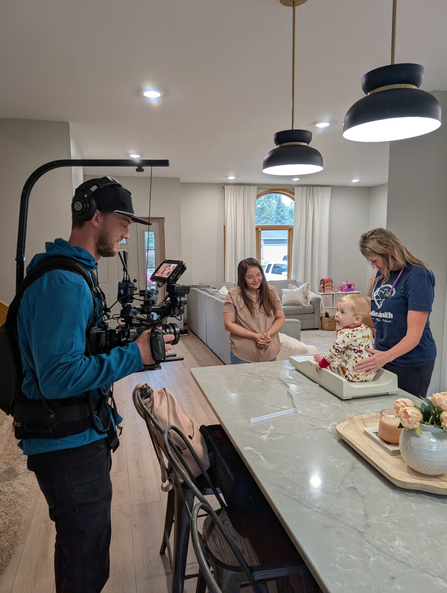 A cinematographer wearing a camera rig films in a bright, modern kitchen. At the counter, a baby sits in a small seat while a woman in a navy shirt stands beside them. Another woman in a light pink top stands nearby, smiling. The kitchen features pendant lights, bar stools, and a vase of flowers on the island, with a cozy living room visible in the background.