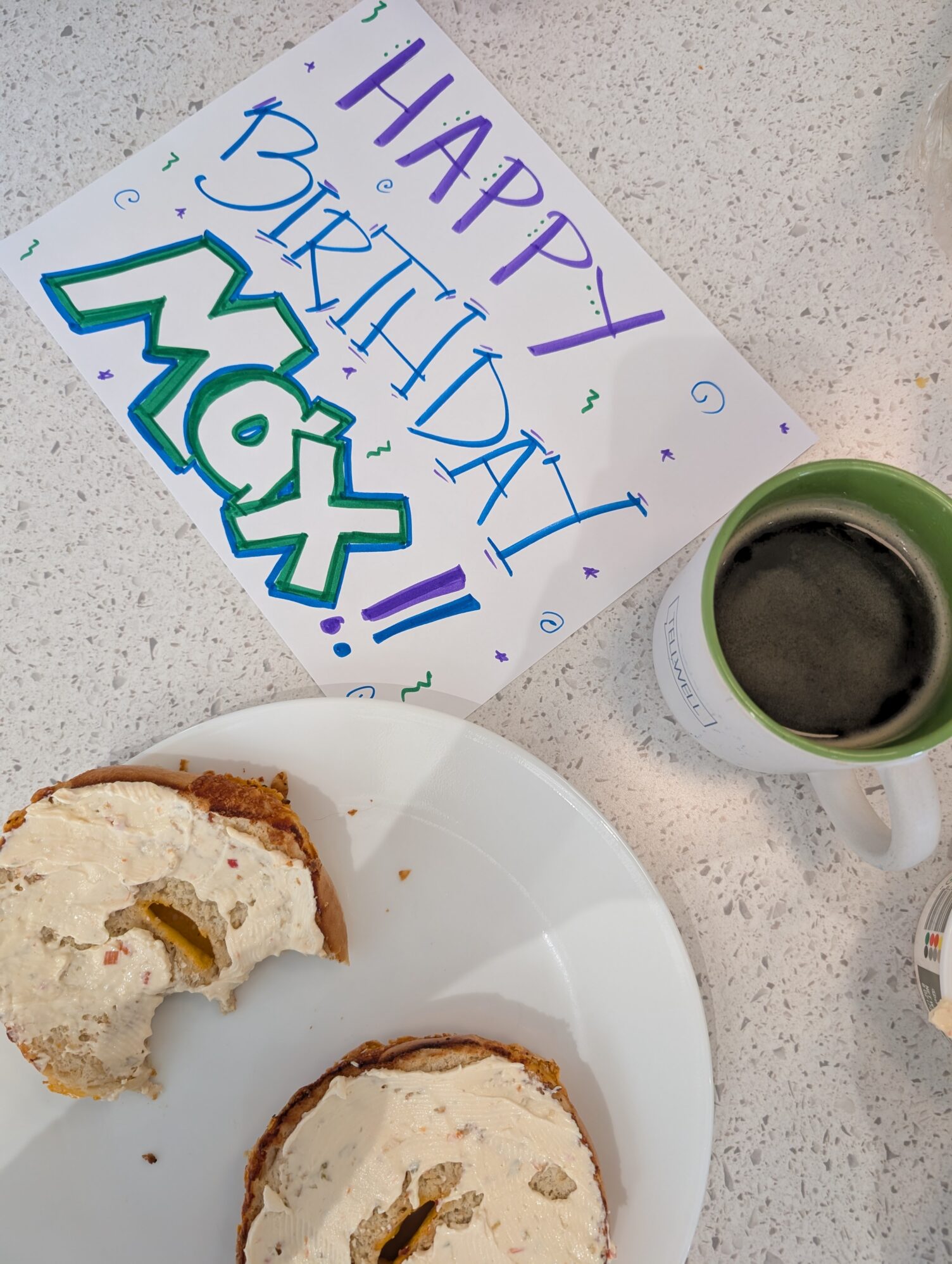 A bagel with cream cheese sits on a plate next to a mug of coffee and a sign that reads “Happy Birthday Max!”