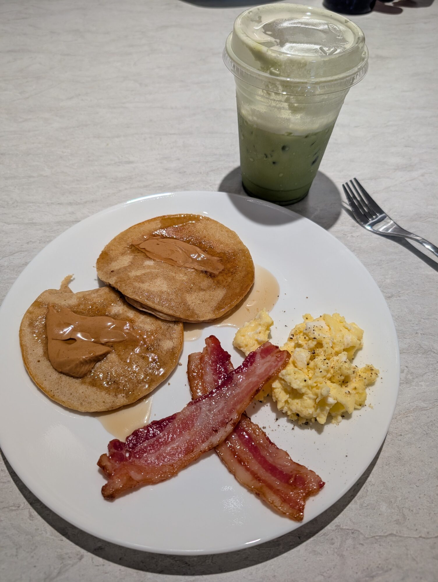 A breakfast plate with pancakes topped with syrup and peanut butter, crispy bacon, and scrambled eggs, with a green iced drink on the side.