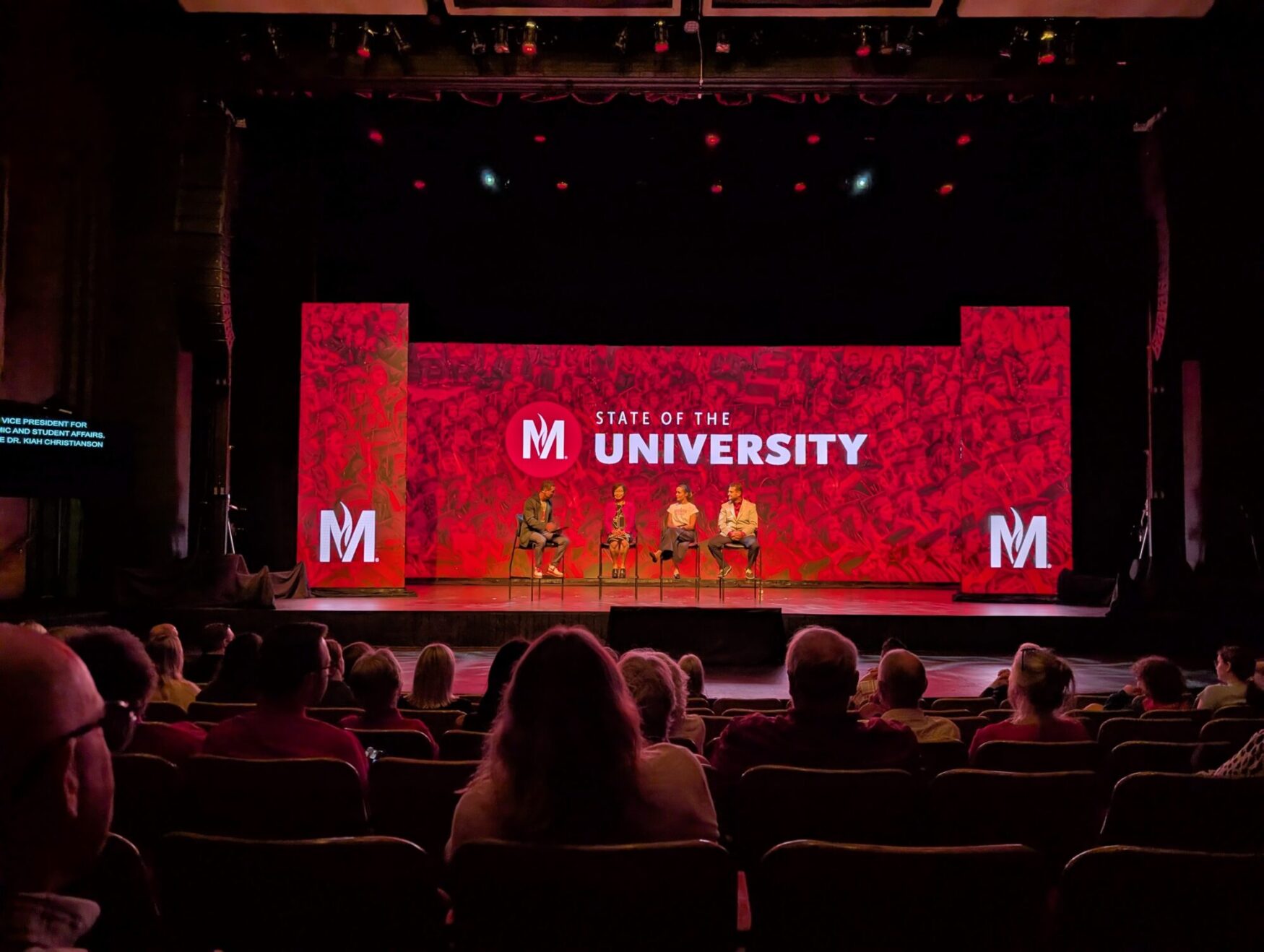 A stage with red electric screens reading “State of the University”. There are four people sitting in chairs on the stage. You can see the audience in the foreground seated in the auditorium.