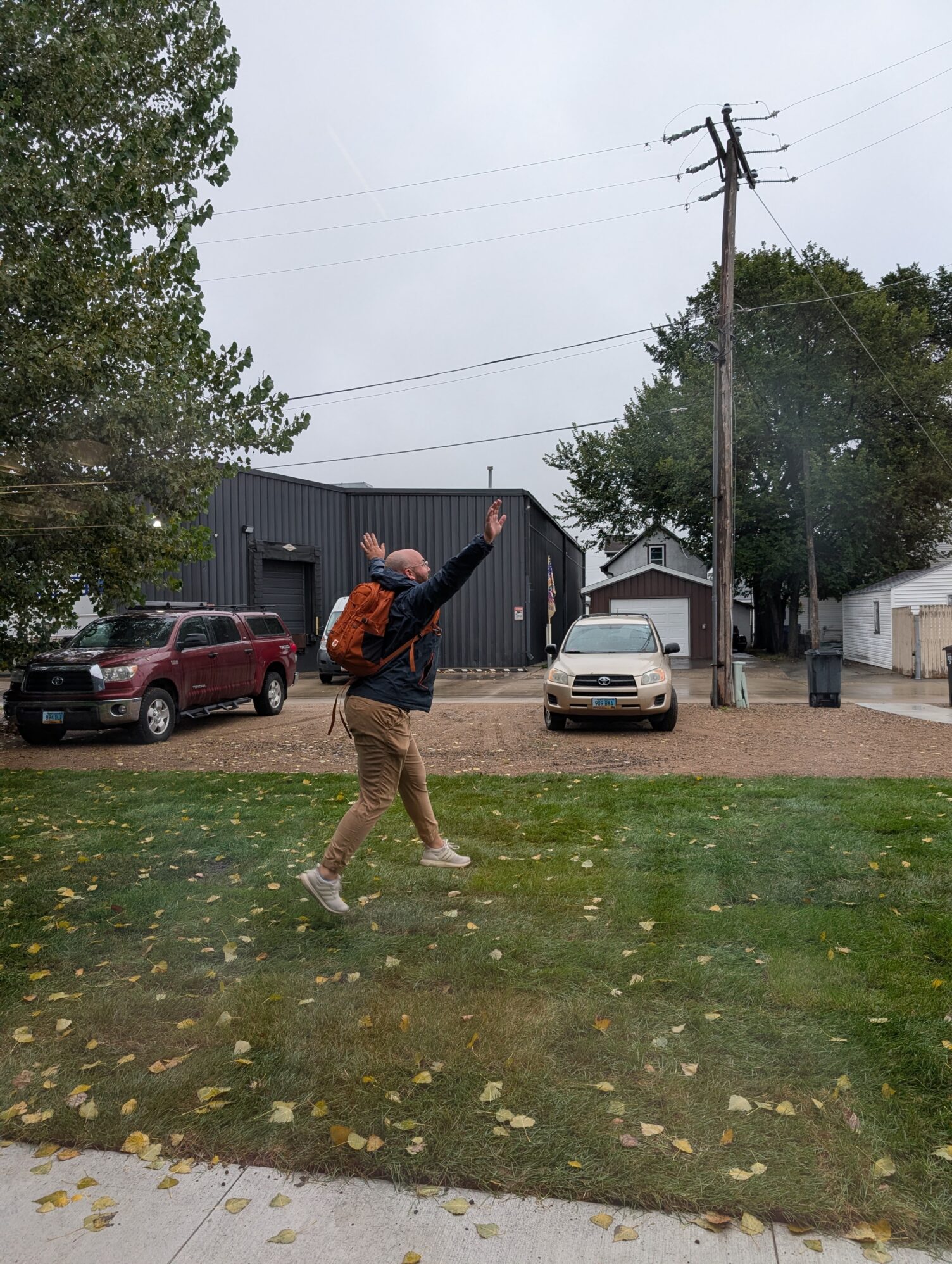 A man wearing a black rain jacket and orange backpack jumps with his arms raised. He is on a lawn with gravel in the background. There are two cars parked on the gravel and a large black warehouse as well.