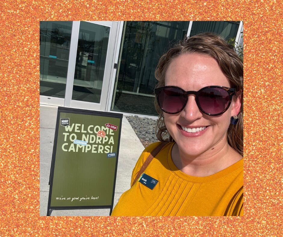 A woman wearing sunglasses and a mustard yellow top smiles for a selfie outside a building. A green sign beside her reads “Welcome to NDRPA Campers! We’re so glad you’re here!”