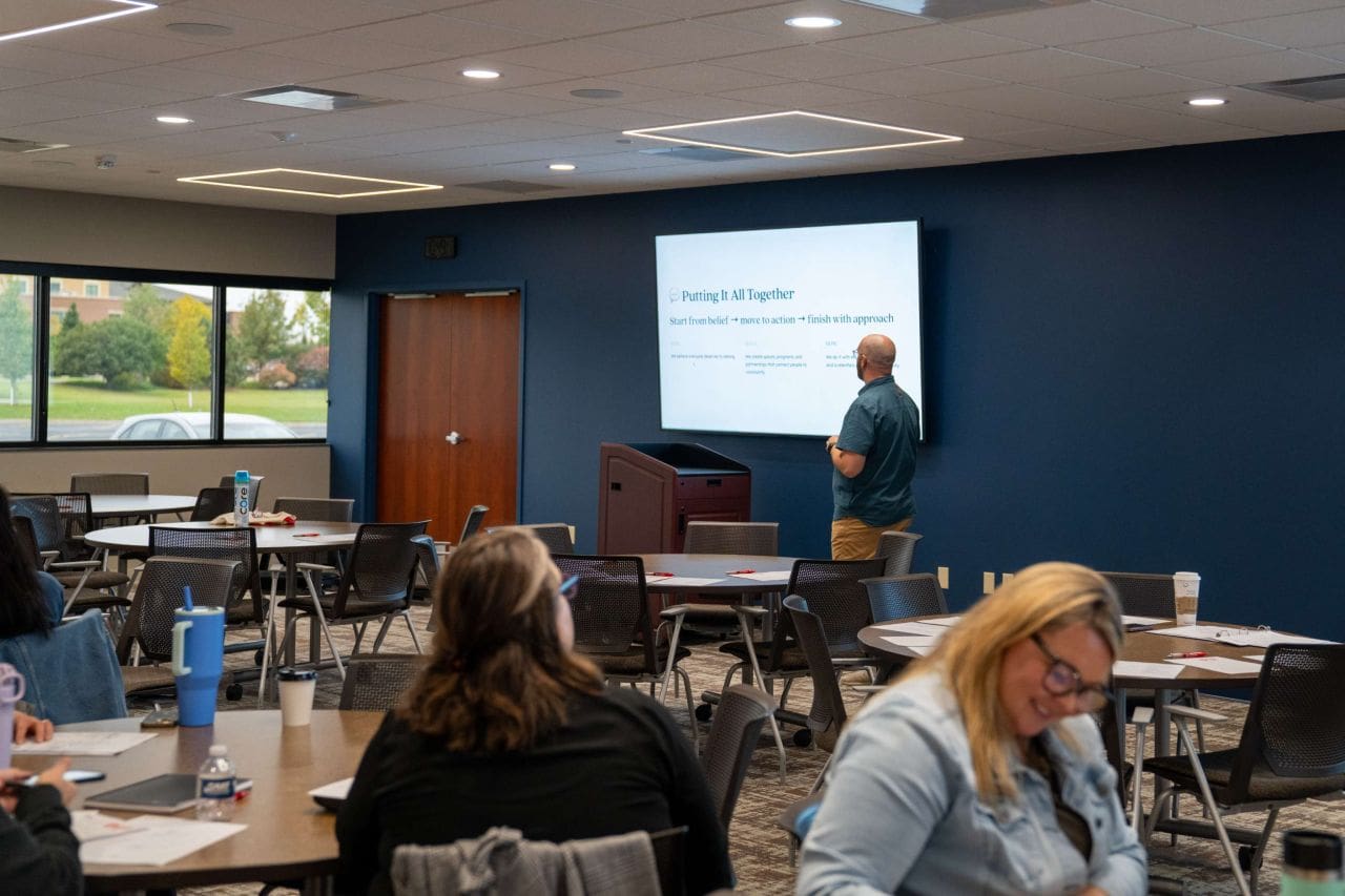 A man in a blue shirt stands in the front of a room next to a projector screen, giving a presentation. There are several people sitting at tables, taking notes while he talks.