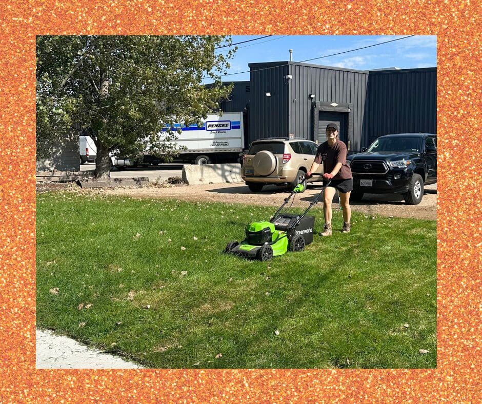 A woman in a dark shirt and shorts pushes a bright green lawnmower across a grassy area outside. Behind him are parked cars, a large tree, and an industrial-style building.