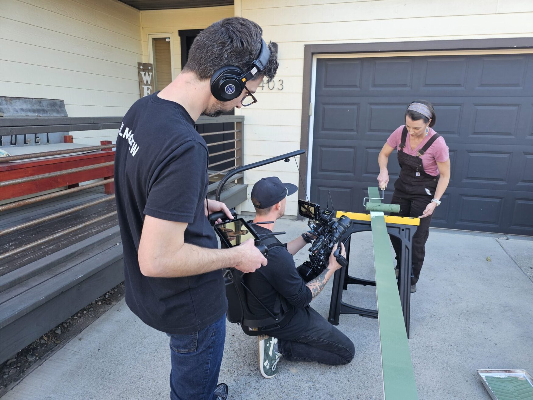 A woman wearing overalls and a pink shirt paints a long green wooden board using a roller outside a garage. Two crew members film her, one holding a camera rig at a low angle and the other wearing headphones and monitoring audio.