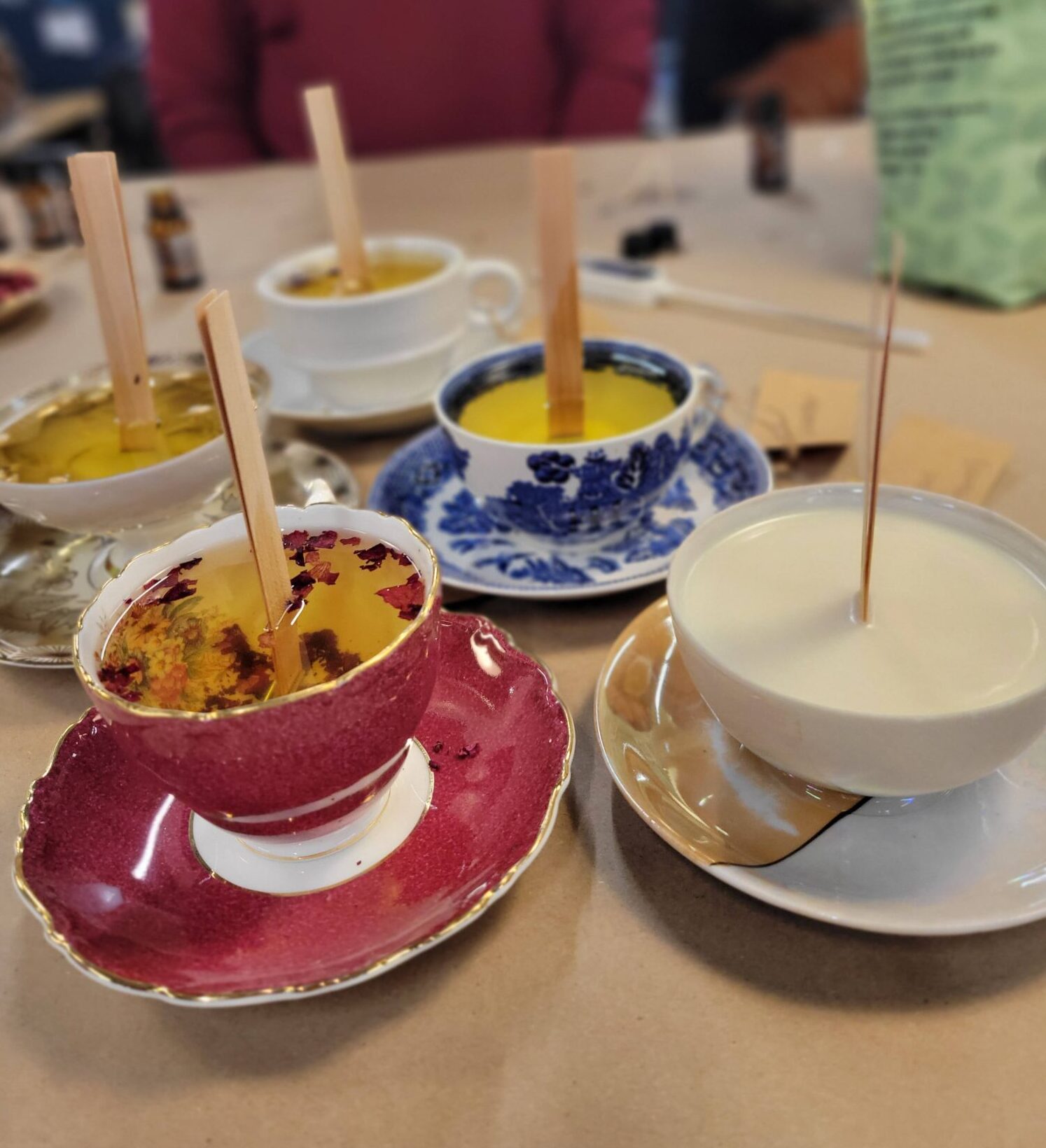 Several teacups filled with wax and wooden wicks are arranged on a table, likely during a candle-making activity. The cups are in different colors and patterns, including red, blue, and white. A person in a maroon sweater is sitting in the background.