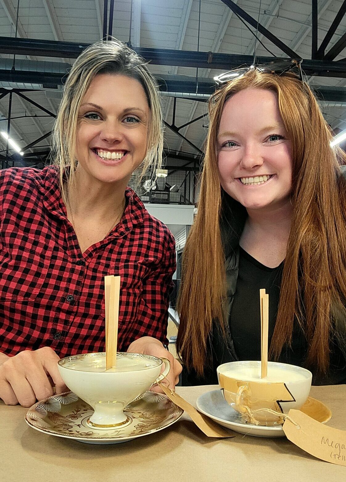 Two smiling women sit at a table holding teacup candles they made. Each candle is in a decorative teacup with a wooden wick and a tag attached. The setting appears to be a workshop or studio with exposed beams and bright lighting overhead.