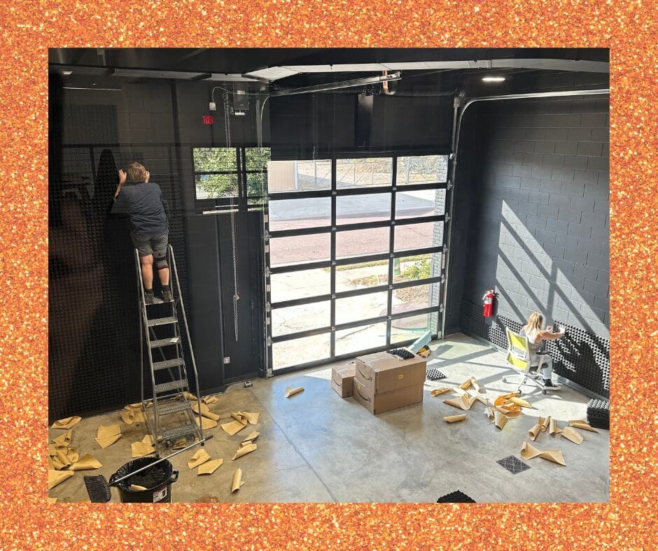 Inside a high-ceilinged black-walled space, one person works on a ladder while another sits on a chair near the wall, attaching black acoustic panels. Cardboard pieces and boxes are scattered across the floor.
