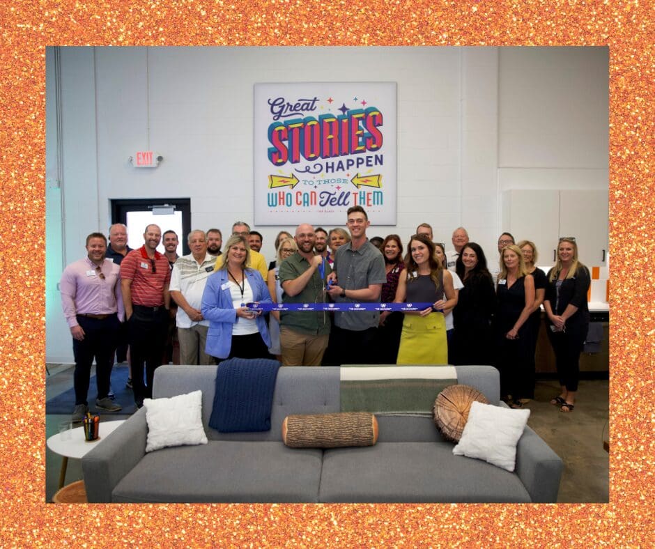 A group of people smiling and standing together at a ribbon-cutting ceremony inside a modern office space. Four individuals in front hold the ribbon, while others gather behind them. A colorful wall sign above reads 'Great stories happen to those who can tell them.' The space features couches, decor, and a kitchenette in the background.