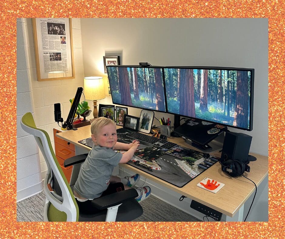 A young child smiles while sitting at a modern workstation with three widescreen monitors showing a forest background. The desk is decorated with family photos, a lamp, and office supplies.