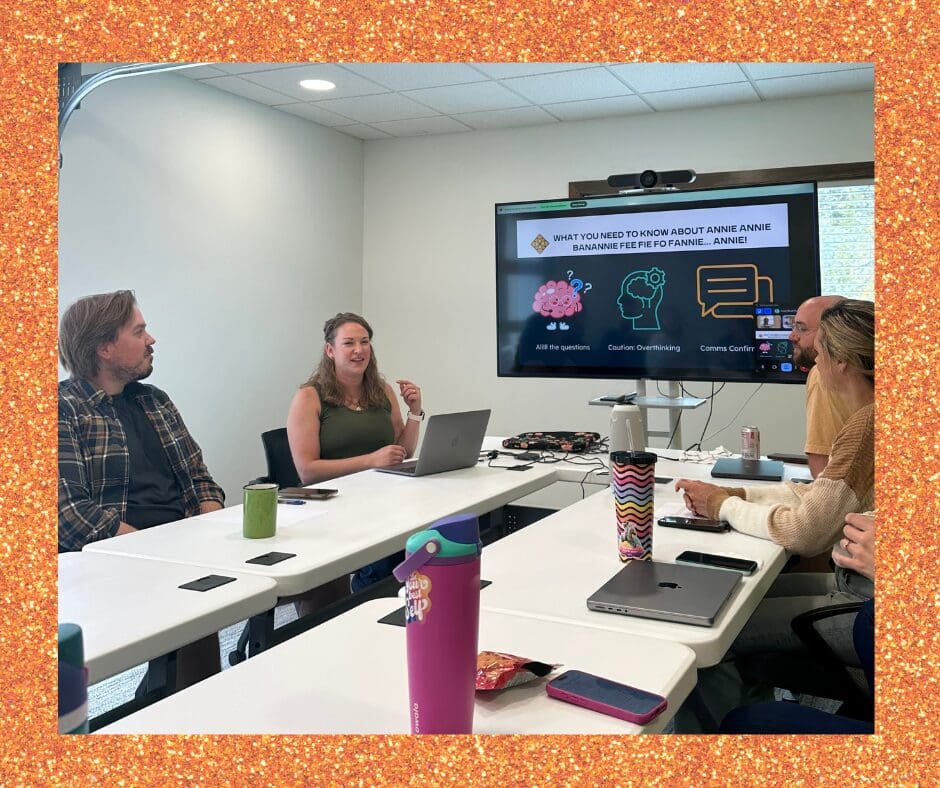 Four people sit around white tables in a conference room. A woman leads the discussion while gesturing beside a large screen that displays text and icons. Others listen with laptops, notebooks, and drinks in front of them.