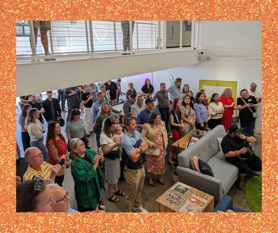 A crowd of people stand in an office space, watching something off-camera. Many hold phones to capture the moment. A staircase and mezzanine level are visible above them.