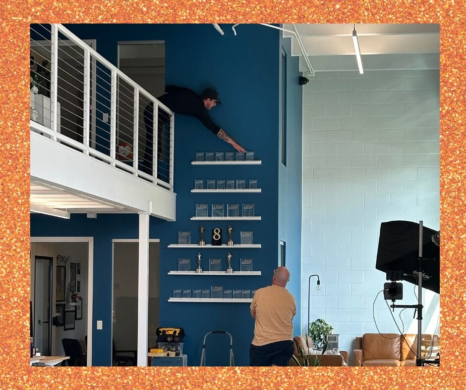 Two people arrange award plaques on a tall blue wall inside a modern office. One person leans out from an upstairs railing to place a plaque on a high shelf, while another stands below watching. The wall is lined with rows of trophies and awards, with a large number '8' displayed among them. A photography light and studio setup are visible in the room.