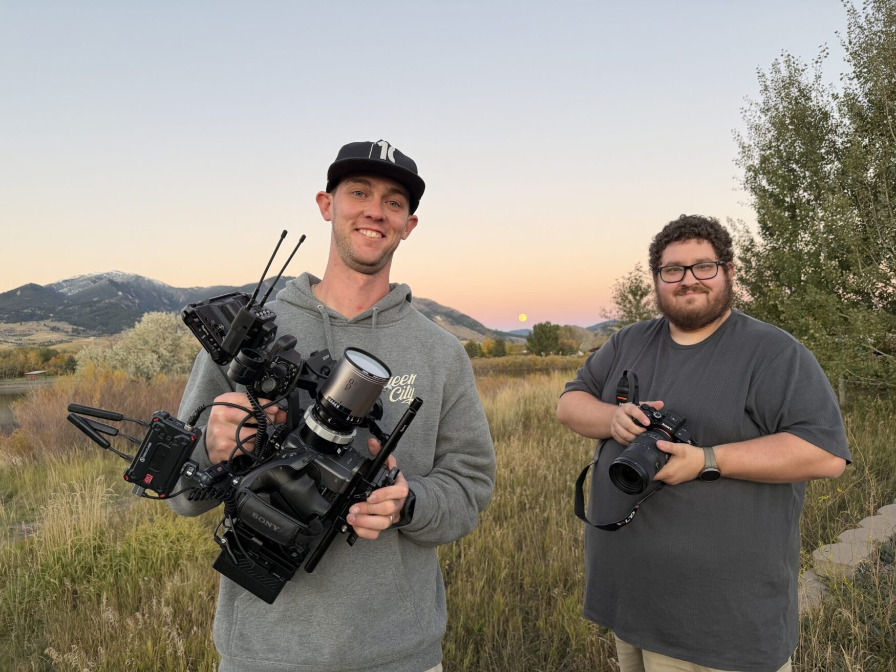 Two men smile for a photo, both holding camera gear. The man on the left holds a professional video camera with additional equipment attached, and the man on the right holds a photography camera. They are in a field at dusk with a mountain range in the background.