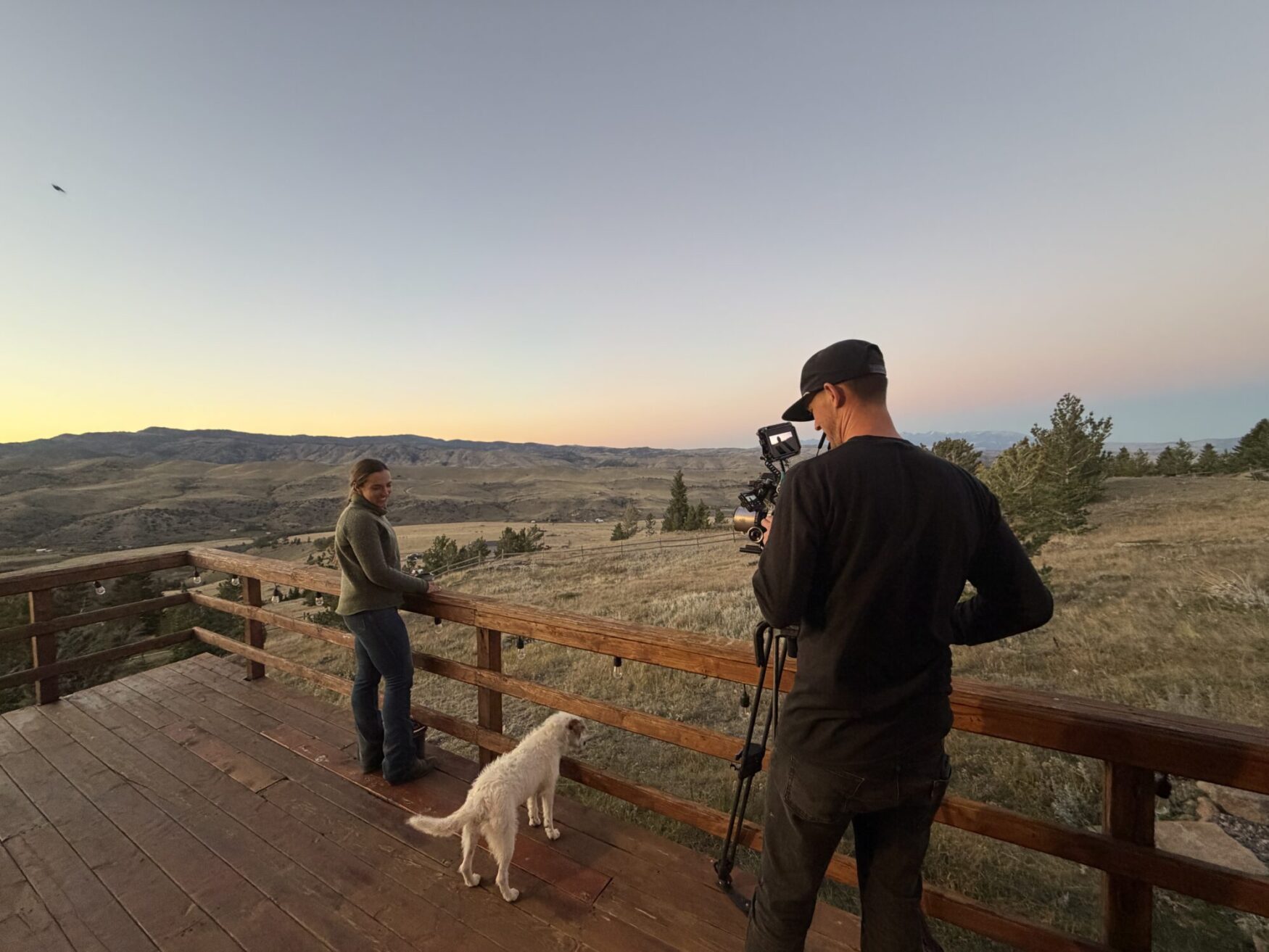 A woman and her white dog stand on a deck overlooking the countryside at dusk. There is a man with a camera on a tripod filming. There are mountains in the background.