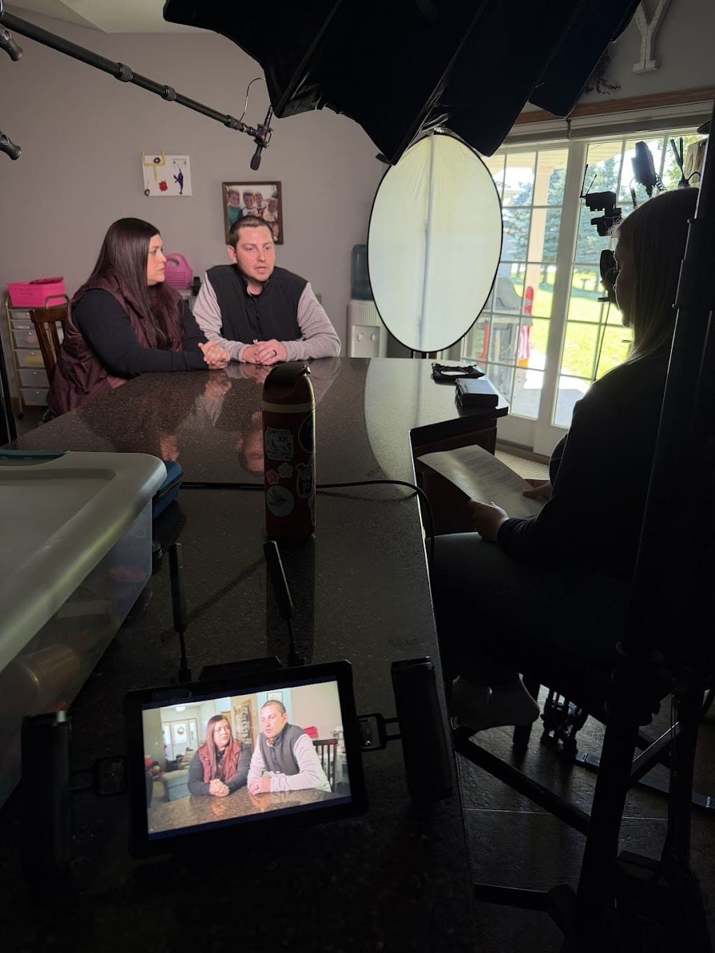 An interview set up in a kitchen. The interviewer is in the foreground, holding a paper with questions. A man and a woman sit next to a microphone while being interviewed. There is a video monitor on the table showing the feed from the camera.