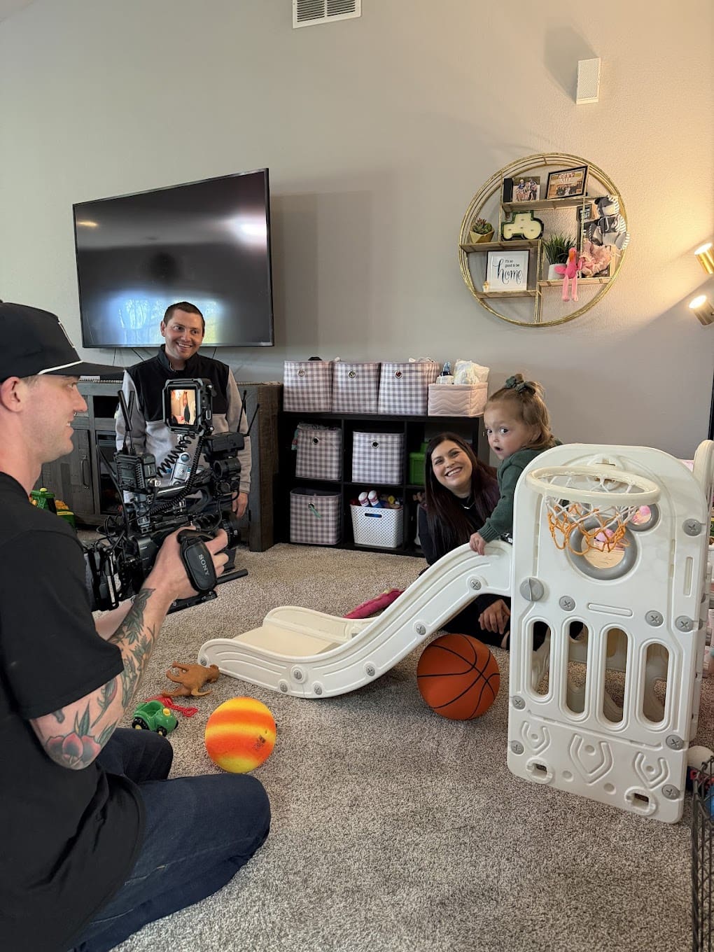 A man and a woman smile at their toddler who is going down a small slide in their living room. The child is looking over her shoulder as a camera man is filming her.