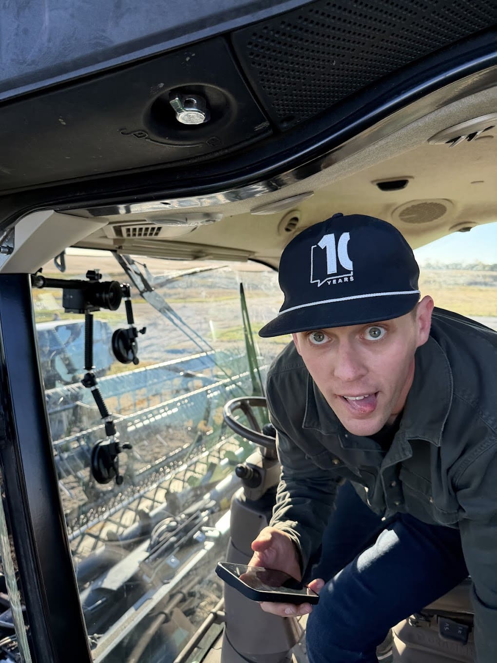 A man wearing a black baseball hat looks at the camera and sticks his tongue out. It appears he is in a farming combine in a field.