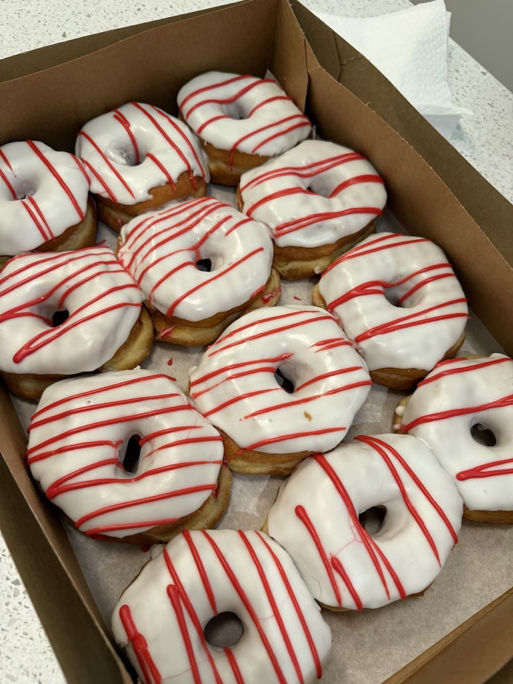 A box of a dozen donuts. The donuts have white frosting with red lines.