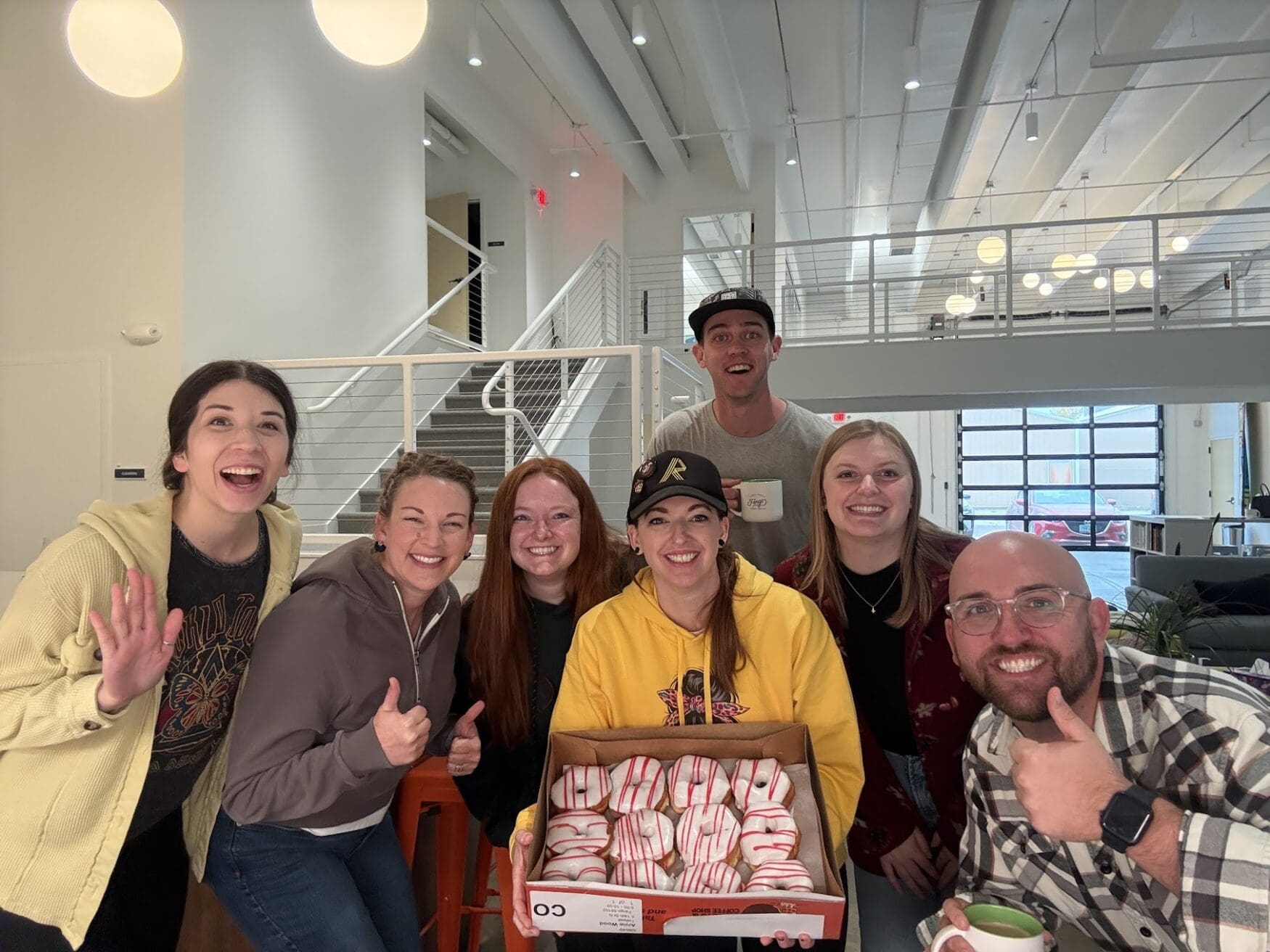 A group of seven people smiling for a photo. Two people are giving thumbs ups while one woman holds a box of red and white donuts.
