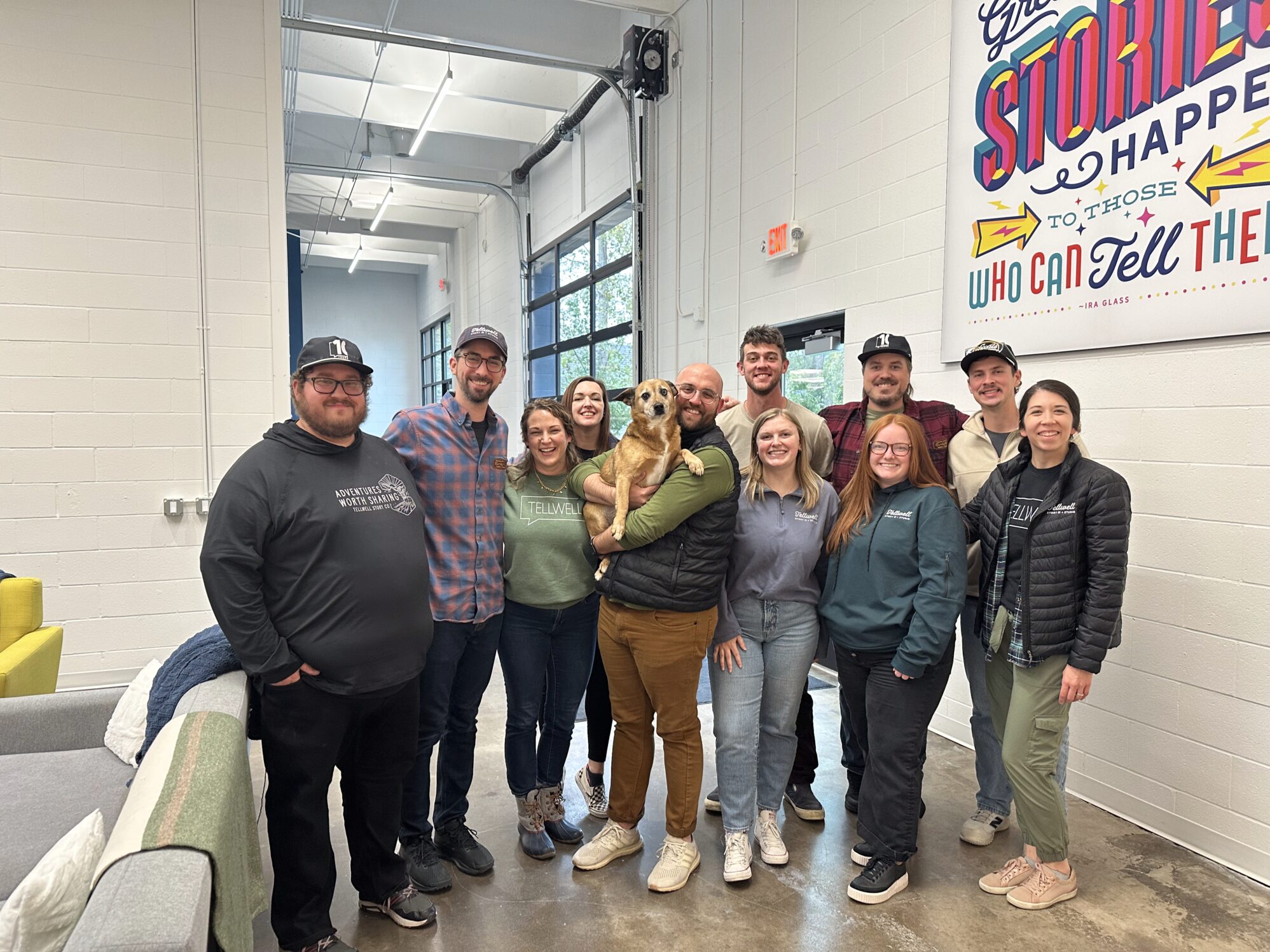 A group of 11 men and women smile at the camera for a photo. They are all wearing a piece of clothing that reads “Tellwell Story Co.” The man in the middle is holding a small brown dog. They are in a large office space with a mural on the right wall.