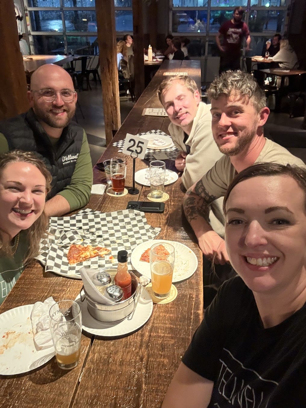 A group of three men and two women smile for a selfie. They are sitting at a large wooden table in a restaurant. There are drinks, plates, and a pizza on the table.