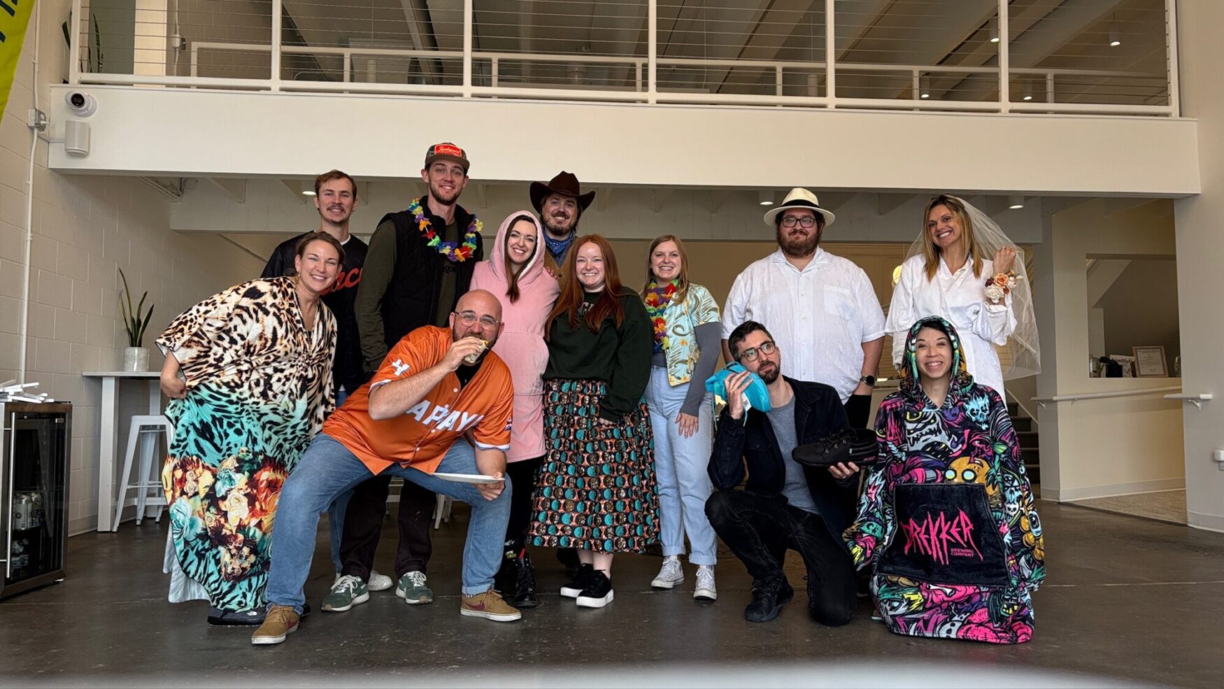 A group of 12 men and women smile at the camera for a photo. They are all wearing fun costumes, including a woman in a bridal veil, a man in a cowboy hat, and more. They are standing in a large, open room with an indoor walkway above them.
