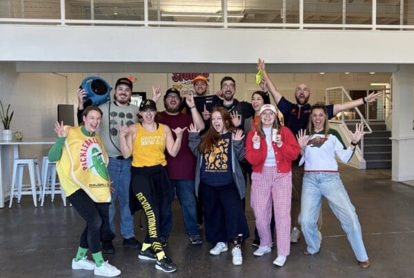 A group of 11 men and women pose for a group photo. They all have excited facial expressions and are wearing festive costumes. They are in a large office space with an indoor walkway behind them.