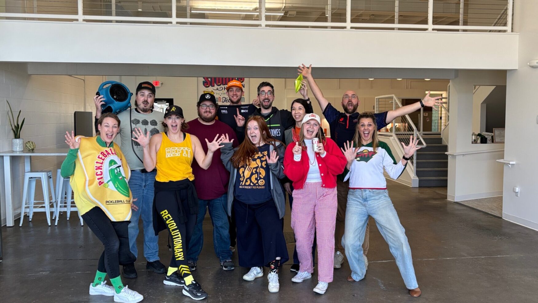 A group of 11 men and women pose for a group photo. They all have excited facial expressions and are wearing festive costumes. They are in a large office space with an indoor walkway behind them.