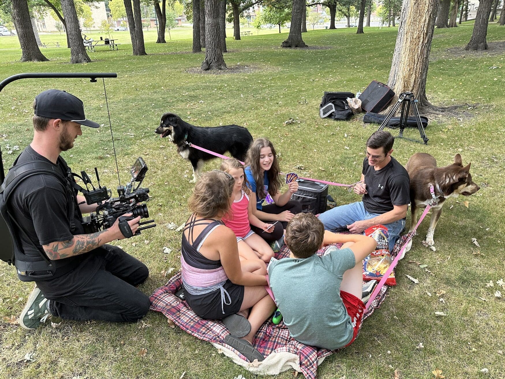 A family of 5 sits on a picnic blanket in the park having conversations and snacks, while a camera man films them. There are 2 dogs on leashes along with additional camera equipment in the background.
