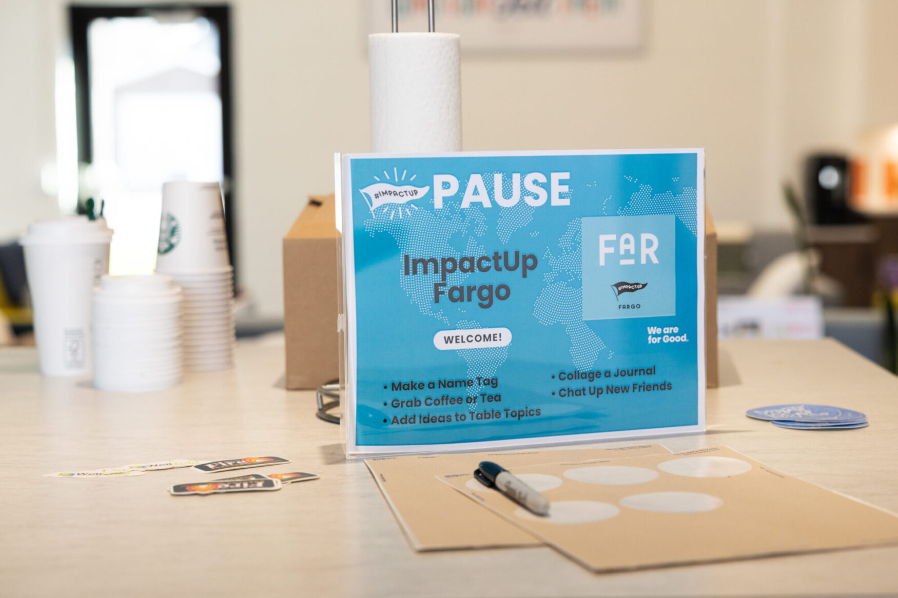 A small poster that reads “ImpactUp: Pause” that is instructing people to fill out a name tag. There are cardboard coffee cups in the background along with stickers and pens.