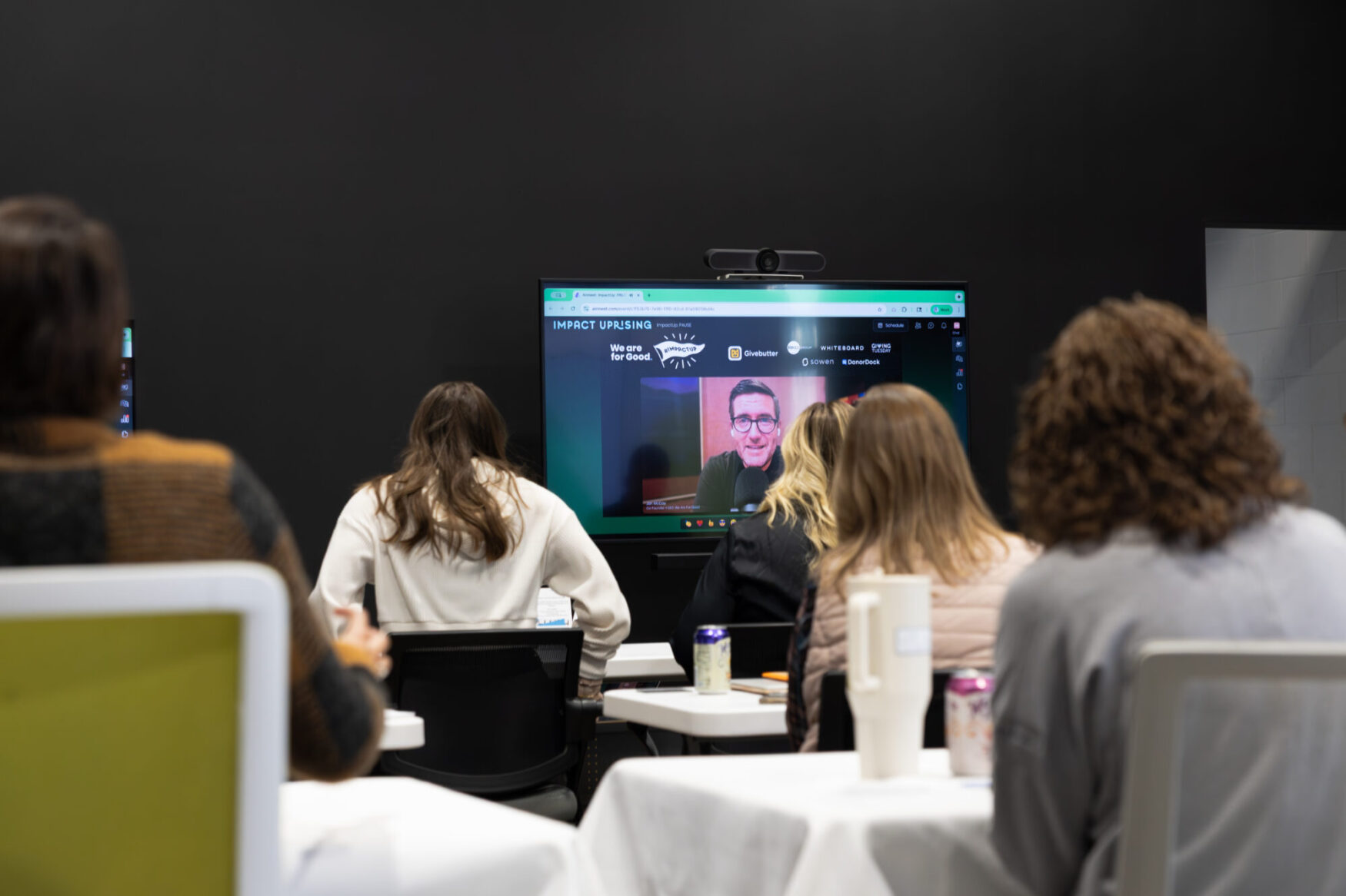 A TV shows a man on a video call with several people in the foreground watching. They are taking notes and listening to the presenter on the screen.