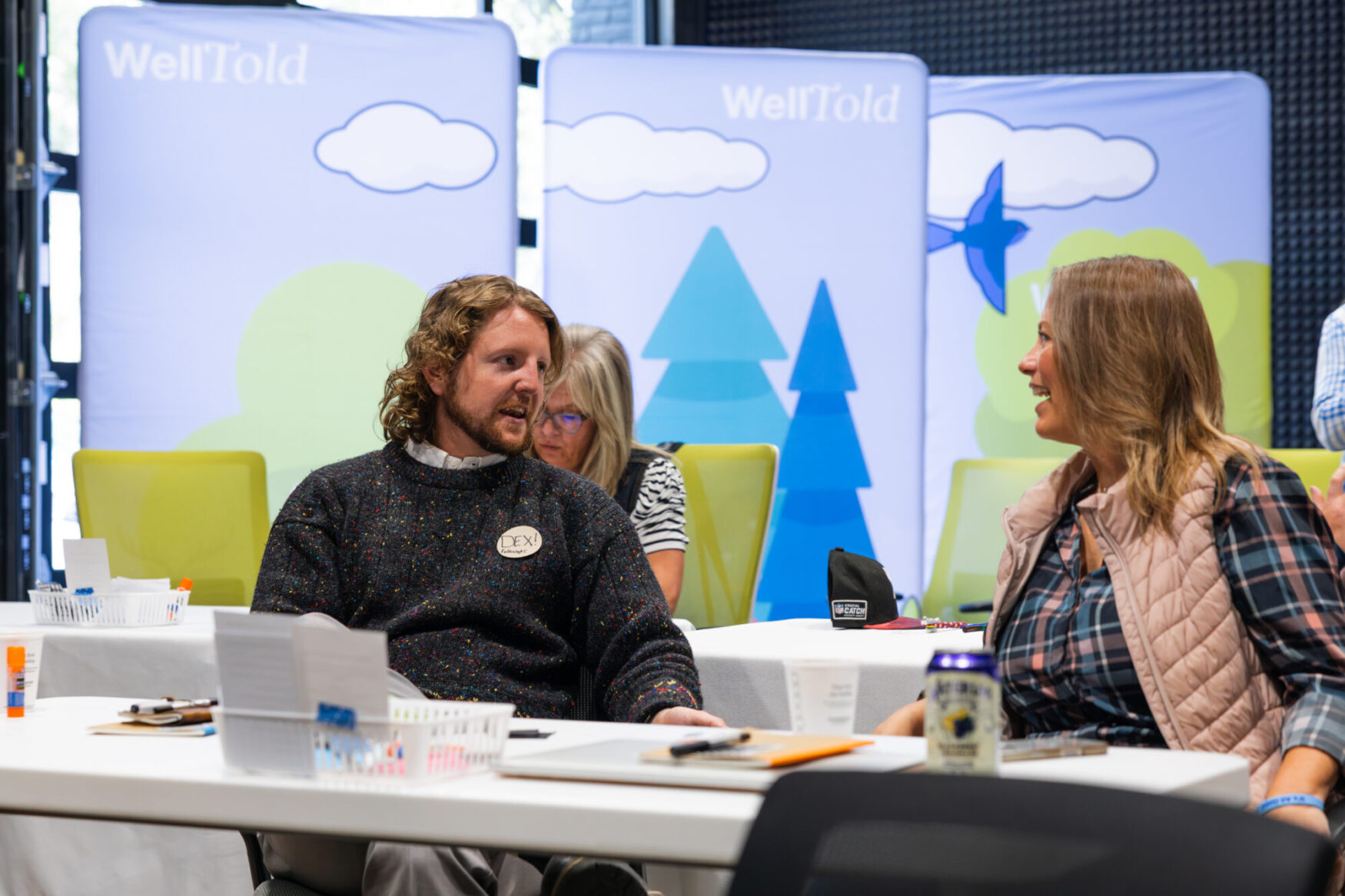 Two people having a conversation at a table. There are laptops, notebooks, and pens on the table. There are three backdrops in the background that read “WellTold” and feature a nature scene.