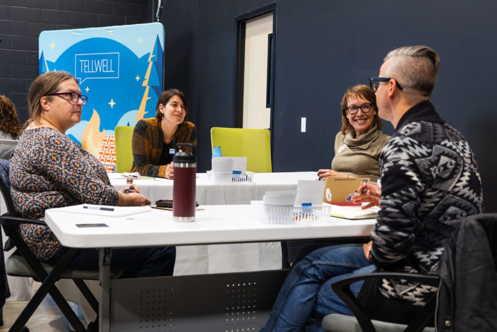 A group of four people seated around a white table. They are in a room with black walls with a backdrop behind them that reads “Tellwell”. They have notebooks and additional work objects on the table and are having a discussion.