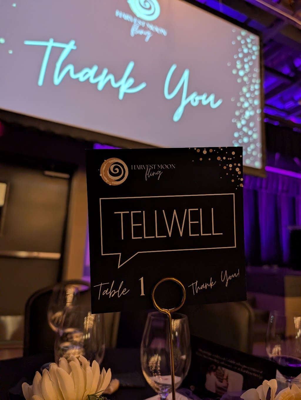 A placecard on a table that reads “Harvest Moon Fling - Tellwell.” There are flowers and wine glasses on the table, as well as a projector screen in the background that reads “Thank you”.