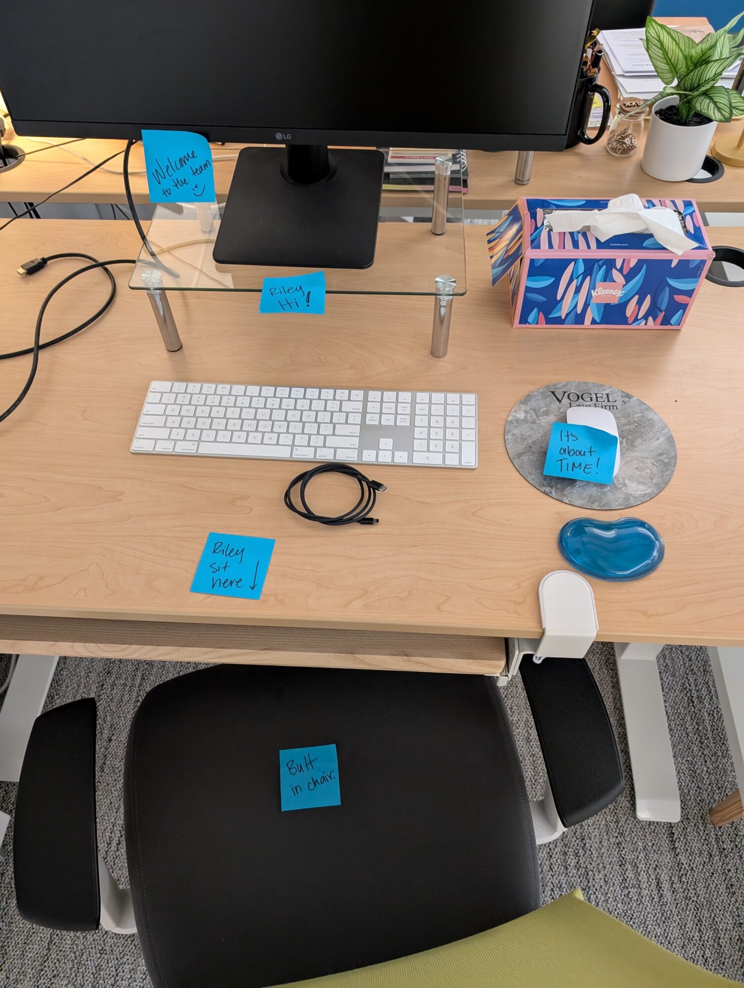 An empty work desk with a computer monitor, keyboard, mouse, and tissue box. There are five post-it notes on the desk and chair, reading “Welcome to the team”, “Riley Hi!”, “It’s about time!”, “Riley sit here”, and “Butt in chair.”