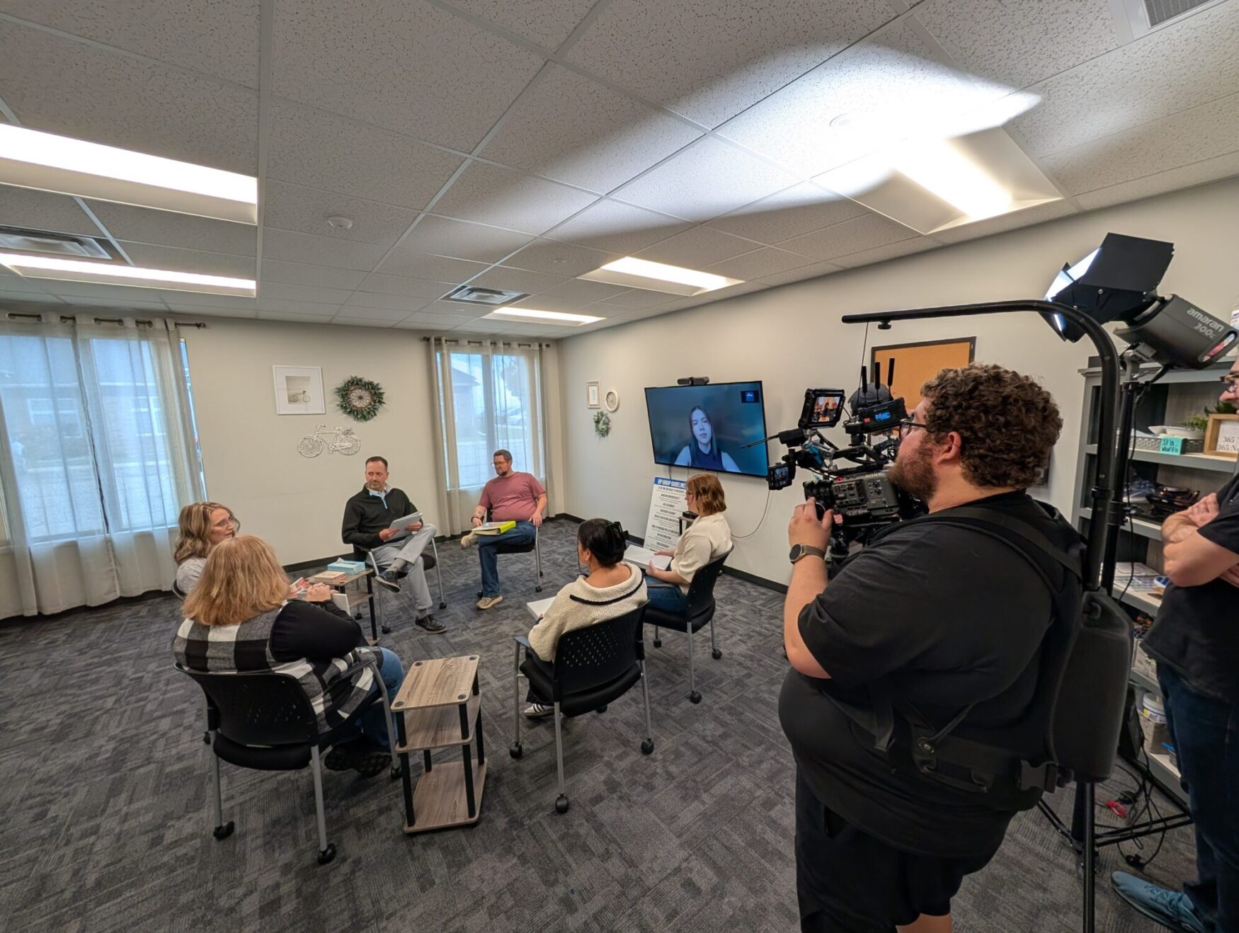 A group of six people sit in a circle in a classroom. There is a man with a camera filming the discussion.