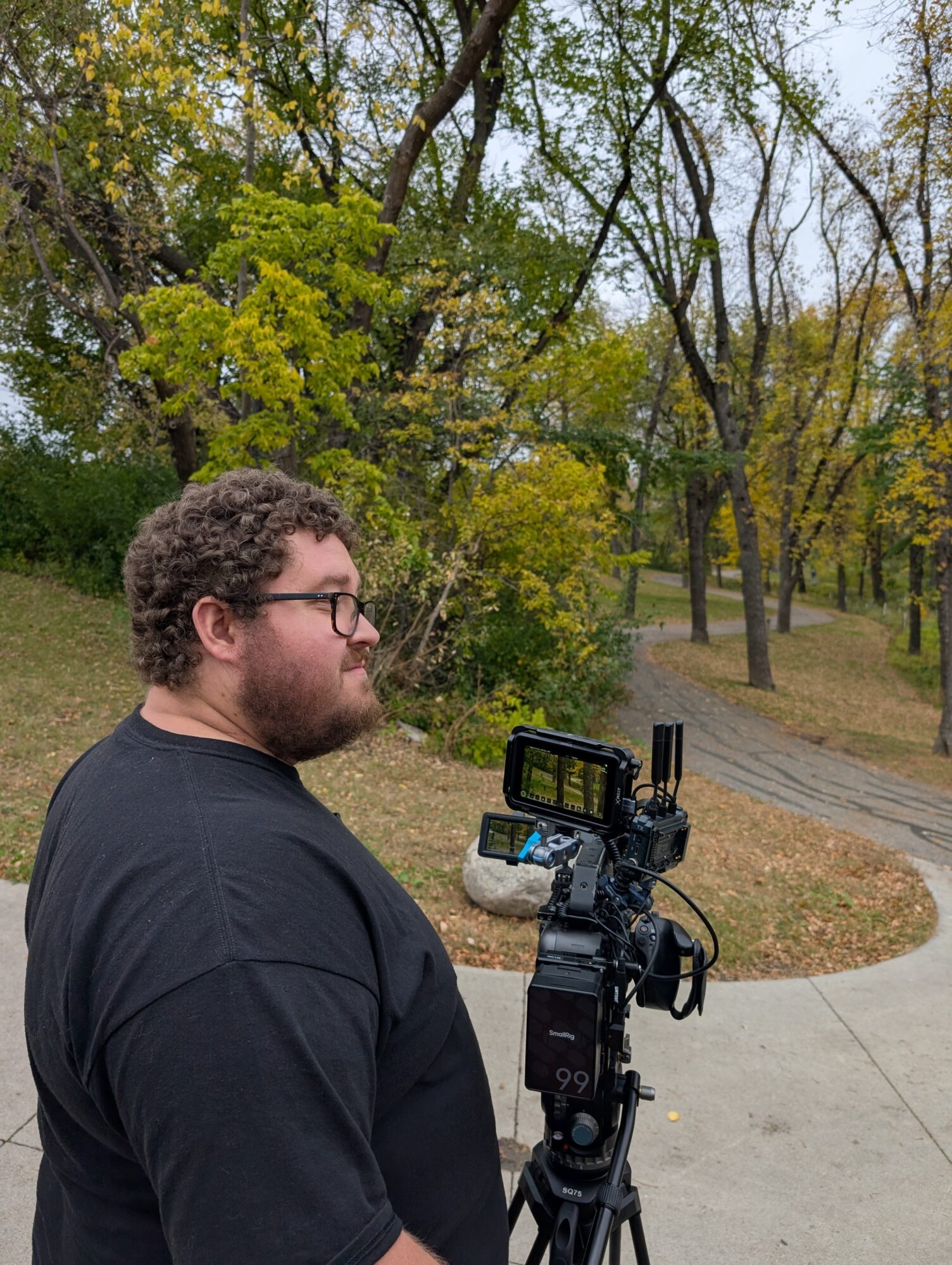 A man with a camera on a tripod looks off to the right. They are in a park with a paved trail.
