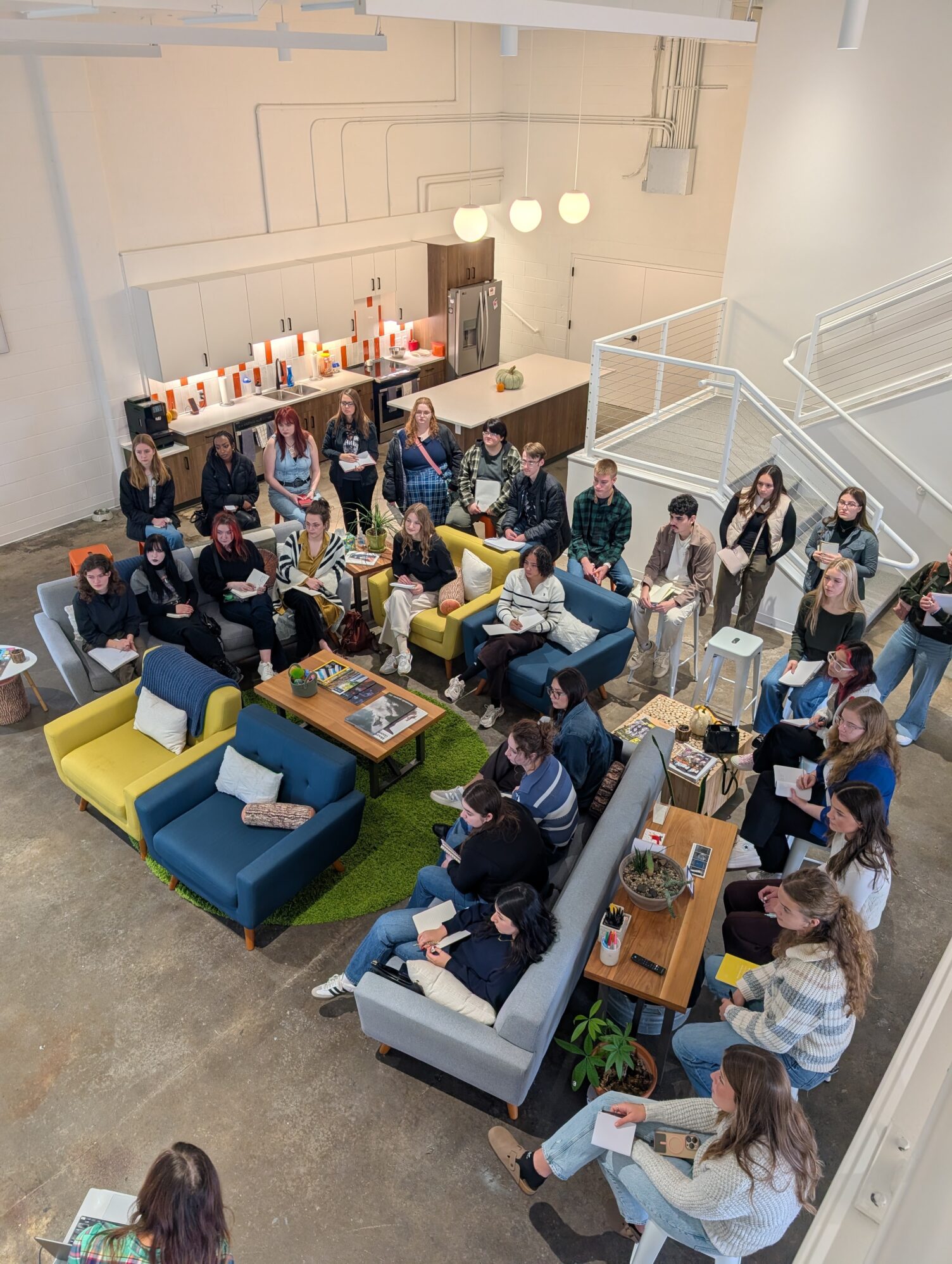 A large group of students sit and stand in a comfortable office setting during a presentation.