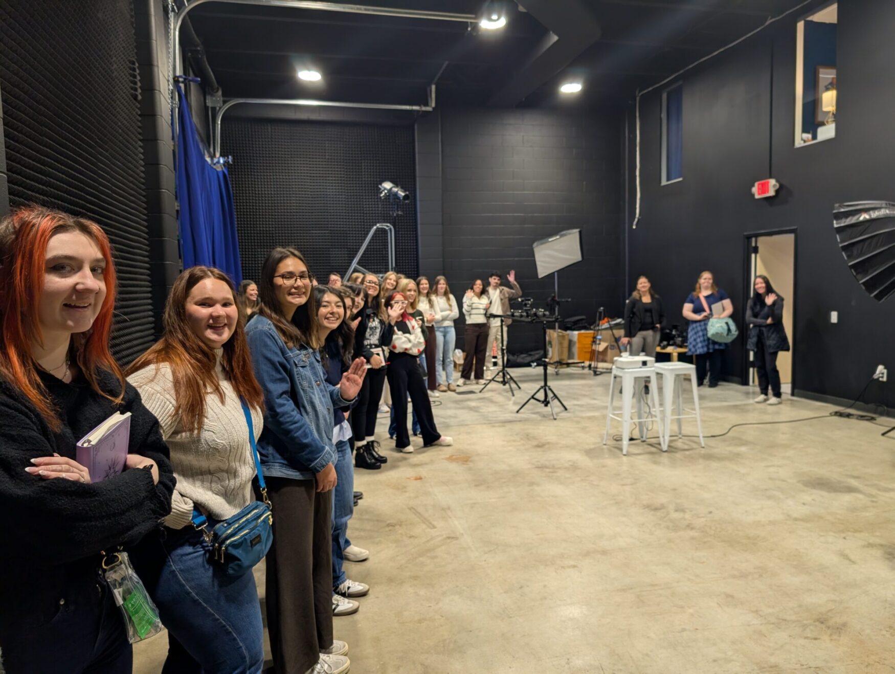 A large group of students stand in a film studio. Some are looking and smiling at the camera. There is camera and lighting equipment throughout the space.