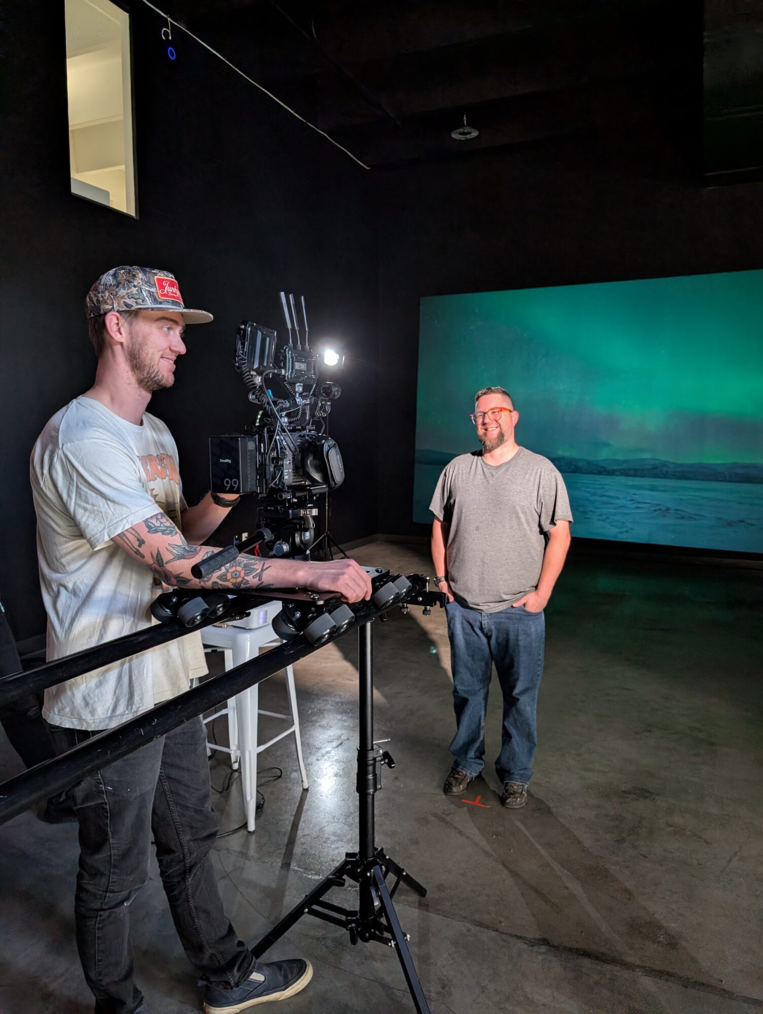 A man with a camera rig smiles as he films another man. There is a projection of the northern lights as the background and they are in a large black film studio.