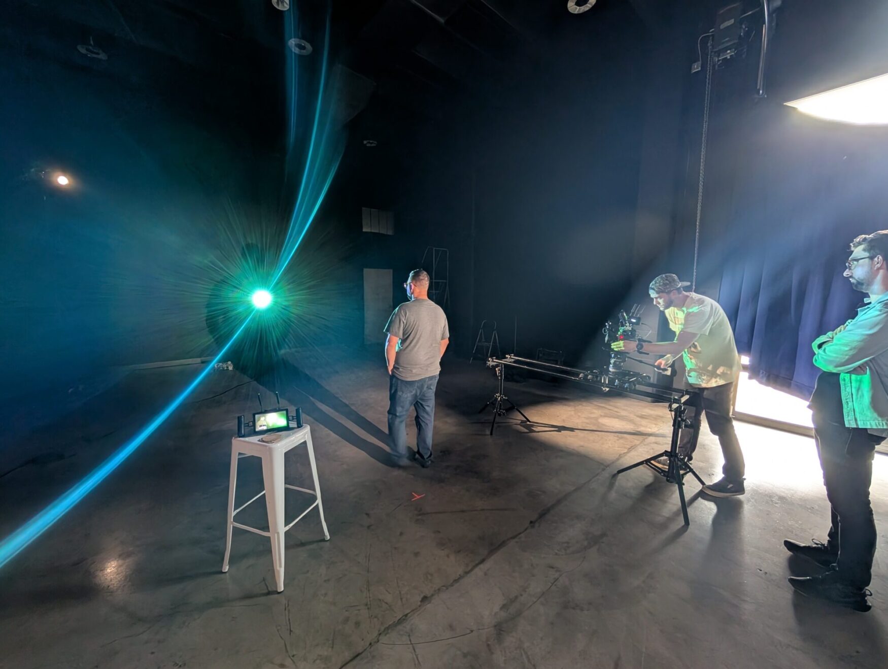 A man with a camera films another man in a large black film studio. There is a projector shining light at the man being filmed. Another man stands to the side and observes.