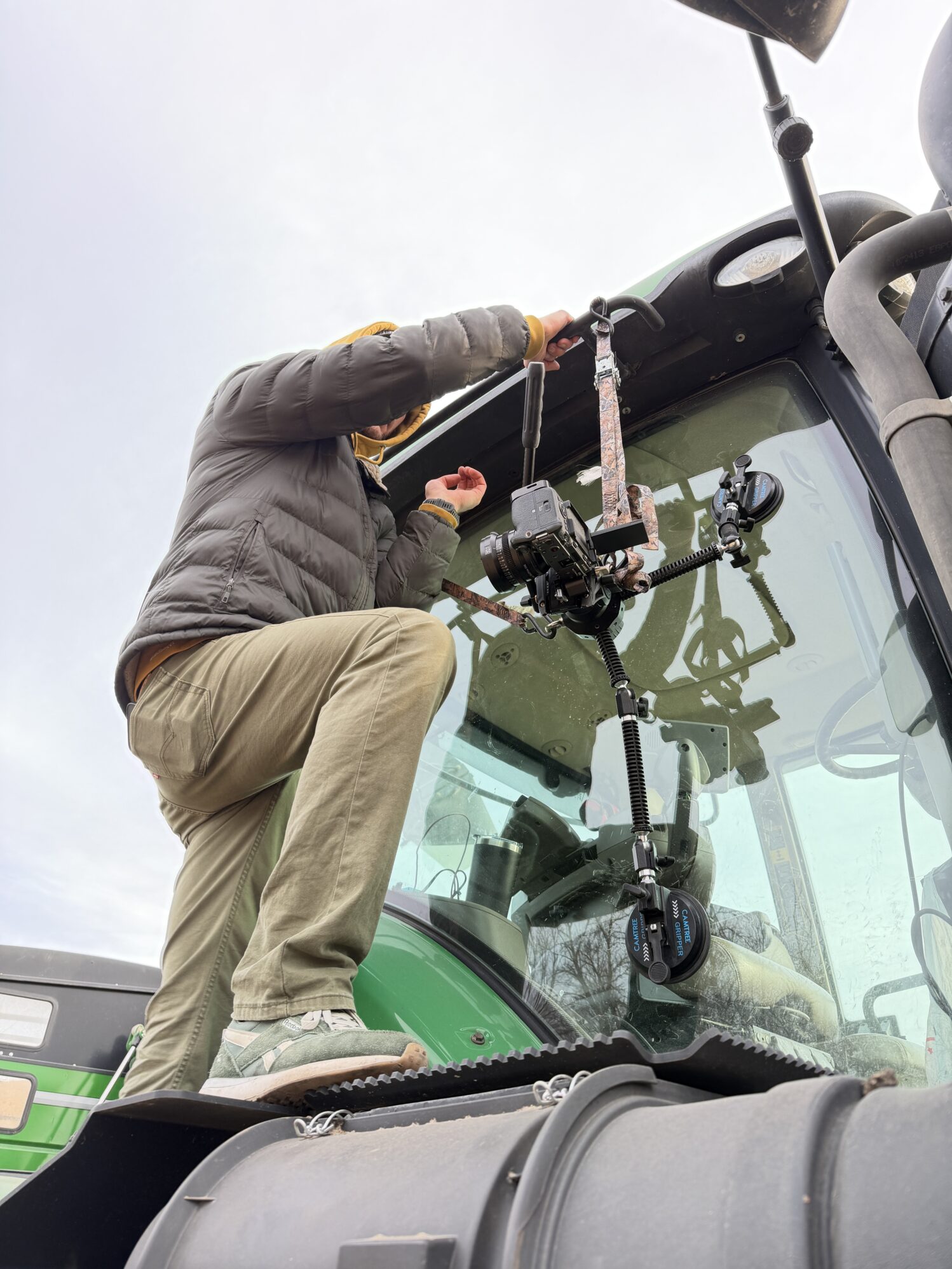 A person standing on the step of a green tractor, adjusting a camera rig attached to the windshield. They’re wearing a gray jacket, khaki pants, and a yellow hood.