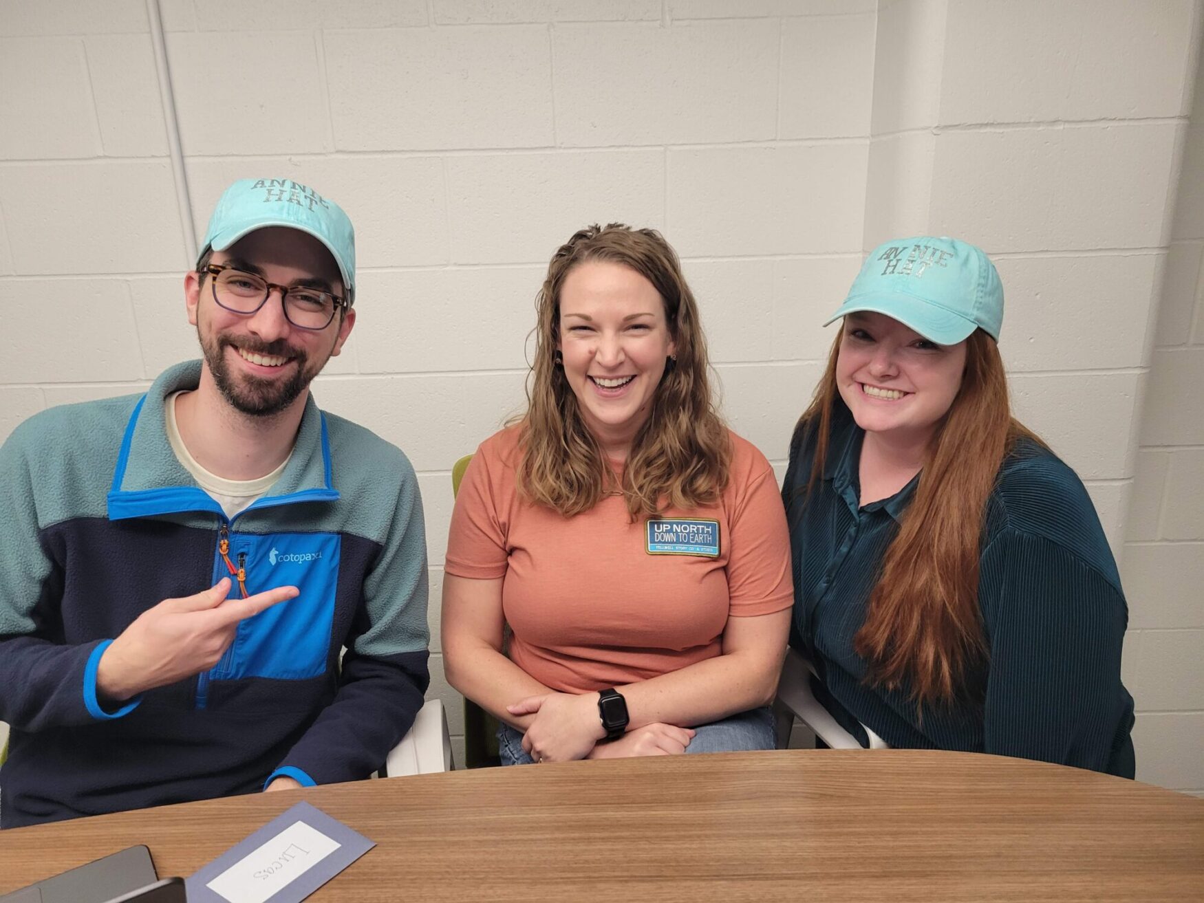 Three people are sitting at a table and smiling at the camera. The person on the left is wearing glasses, a blue fleece, and a teal hat, pointing toward the woman in the middle. The woman in the center is wearing a rust-colored shirt, and the woman on the right has long red hair, a teal shirt, and a teal hat.