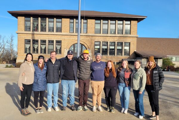 A group of ten people stands smiling in front of a large, historic brick building under a clear blue sky. The mood is friendly and relaxed.