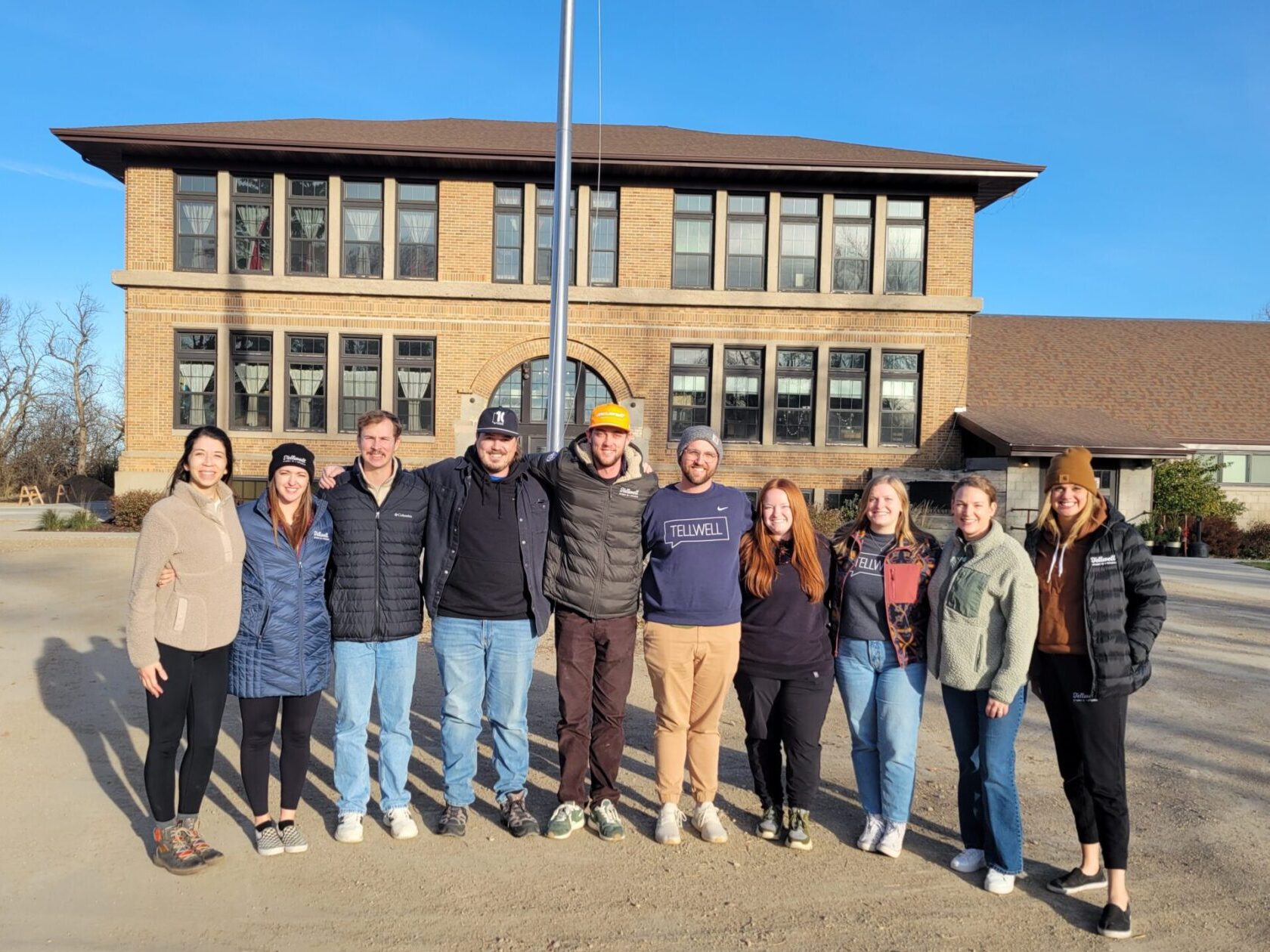 A group of ten people stands smiling in front of a large, historic brick building under a clear blue sky. The mood is friendly and relaxed.